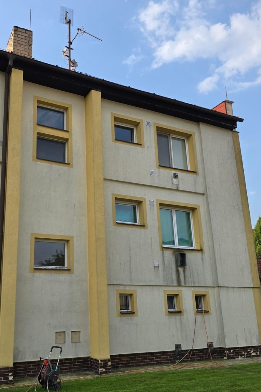 Vícepatrová omítková fasáda se žlutými rámy oken, celoplošně zašedlá s tmavými šmouhami stékajícími od střechy a kolem oken