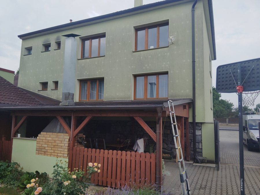Rohový dům se štítem se zelenou omítkou se stopami po dešti a nerovnoměrným šedým znečištěním, dřevěný přístřešek s terasou